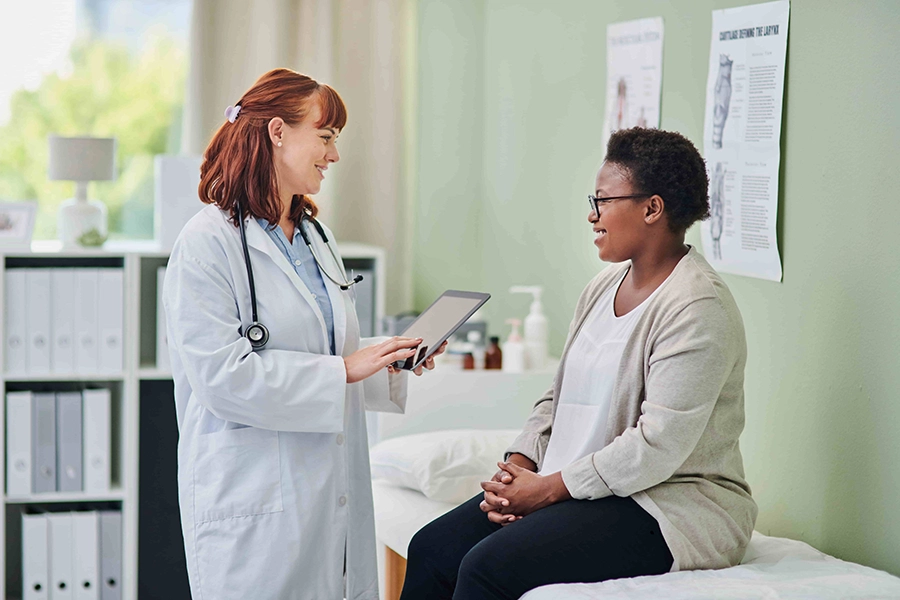 Doctor using a tablet while talking with a patient sitting on an exam table in a clinic exam room