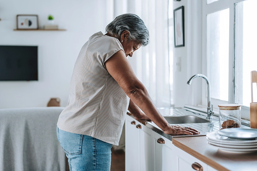Older woman leaning on her kitchen counter with her head lowered, steadying herself with both hands.